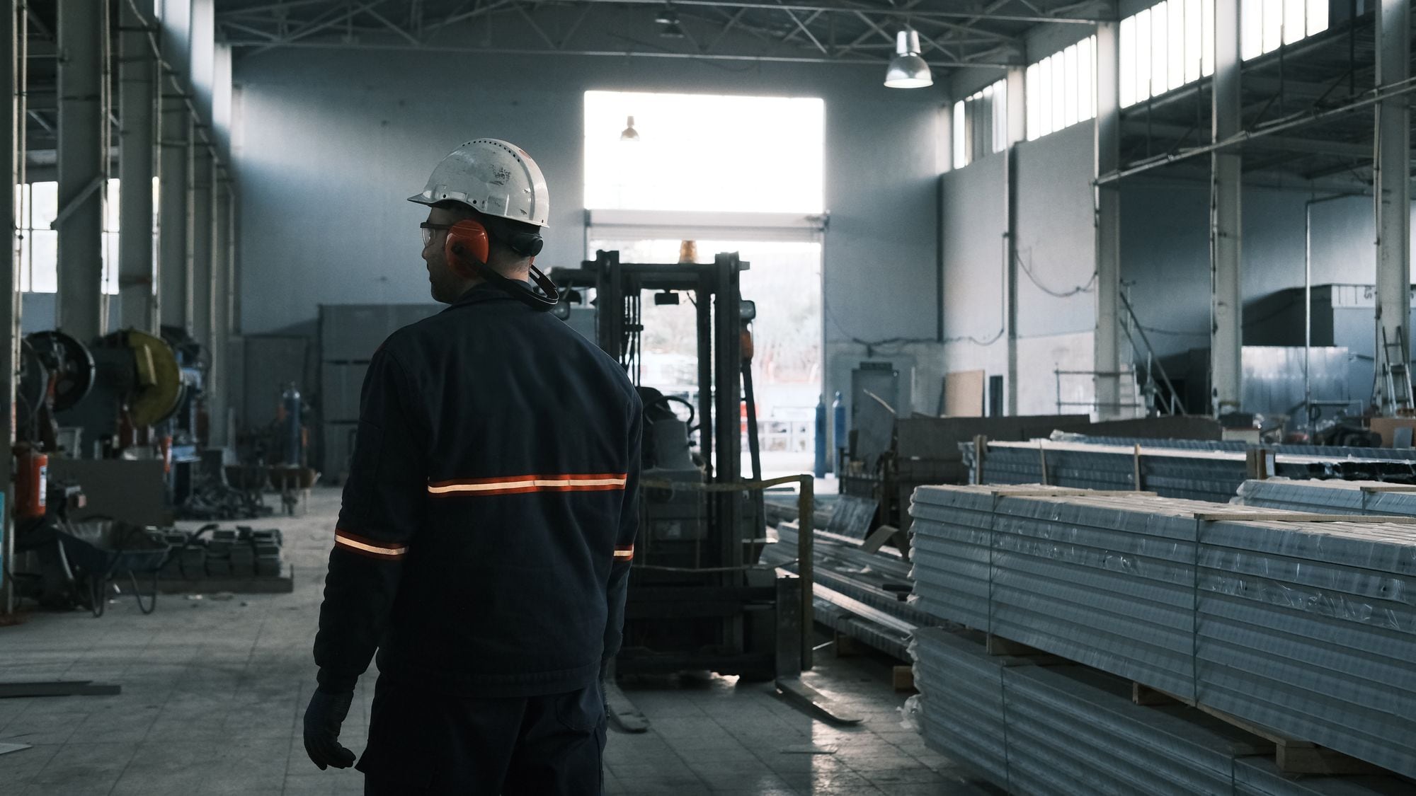 A shop worker in a hard hat and safety earmuffs walks through a manufacturing floor.